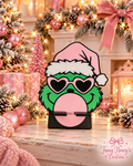 Grinch hair tie holder with pink Santa hat, heart-shaped glasses and bubblegum detail, with two black front arms and a black freestanding base.